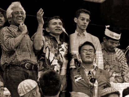 Group of men on a stage at a rally; one man speaks into a microphone while others cheer and raise hands.
